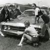 Students in 1970s fashion attire place a bumpersticker on a car. Click for details
