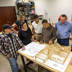 Instructor and students around a table discussing a wood model of a building and blueprints