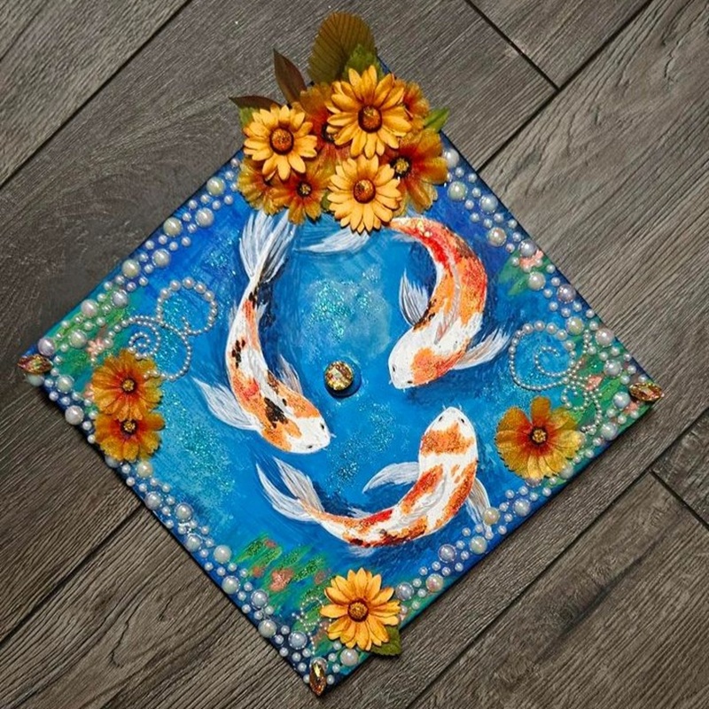 Graduation cap decorated with three koi fish swimming in a circle on a vibrant blue background, adorned with sunflowers, pearls, and golden embellishments.