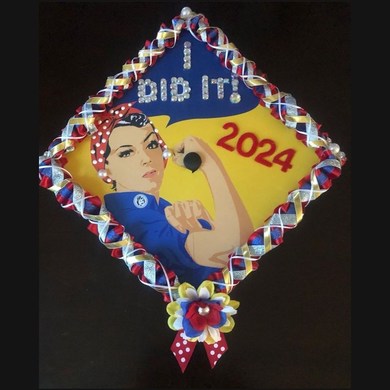 Graduation cap with a design featuring Rosie the Riveter, labeled "I Did It! 2024." The cap is adorned with multicolored ribbons and a floral accent at the bottom.