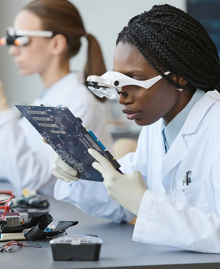 A woman in a lab coat wearing magnifying headgear examines a circuit board