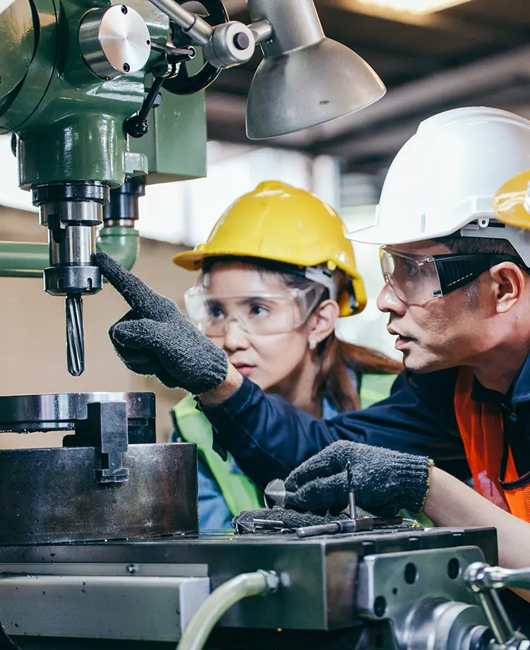 And instructor and student in hard hats and safety goggles inspect a drill press