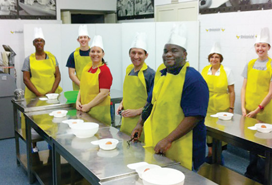 Students in aprons pose in front of thier kitchen work stations