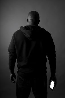 A black man, dressed in a black hoodie, holding a cell phone, standing with his back to the camera.
