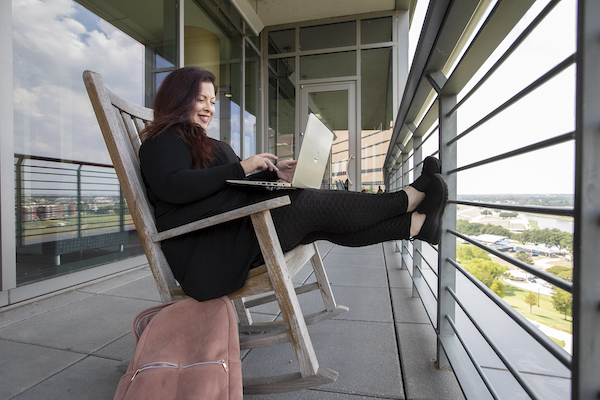 Katie sits on a rocking chair on a patio, working on her latop