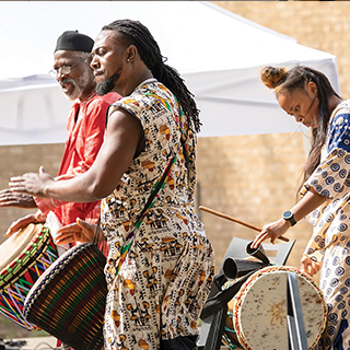 Two men and a woman dressed in traditional African clothing and playing drums.