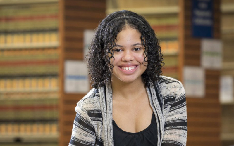 A female student with a dark , curly hair smiles in the foreground. The soft focus background appears to be a library