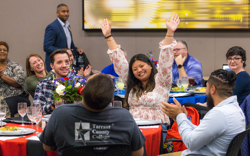 A female professor raises her arms triumphantly above her head while seated at a table. Other faculty and staff can be seen seated around her.