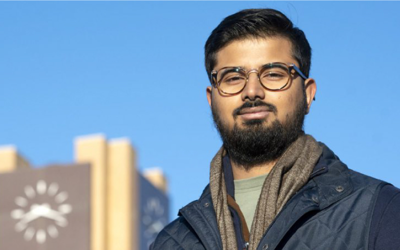 A male student with a dark beard and glasses smiles in the foreground, while the TCC Northeast Campus clock is in the background