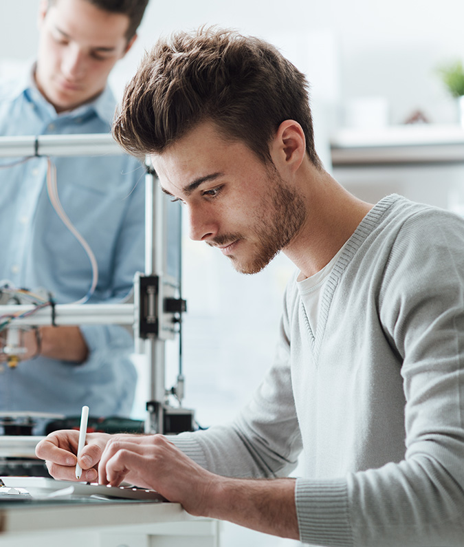 A young man works on a tablet computer while in the background an out of focus man uses a 3D printer. The man in the foreground is learning job skills he can take to the workforce today